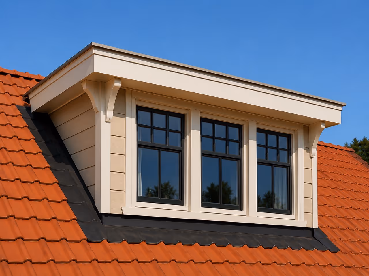 Grote dakkapel met drie ramen en crèmekleurige lijst op rood dak met blauwe lucht.