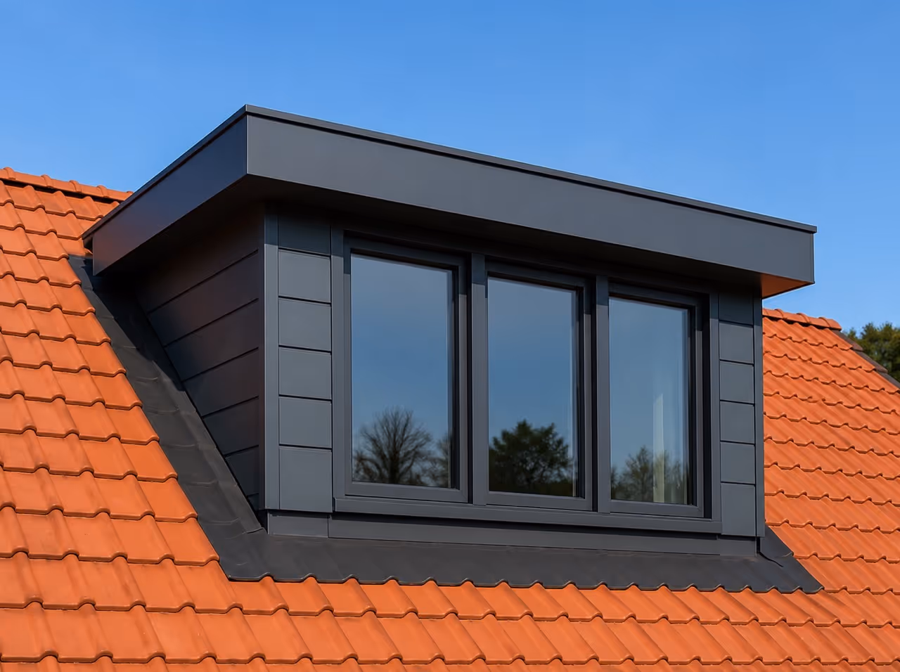 Moderne dakkapel met zwarte bekleding en grote ramen op rode dakpannen onder een blauwe lucht.