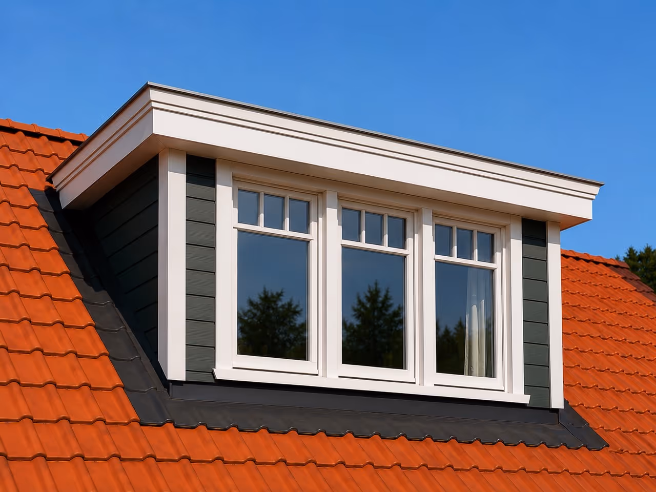 Traditionele dakkapel met witte kozijnen en drie ramen op rood dak tegen blauwe lucht.