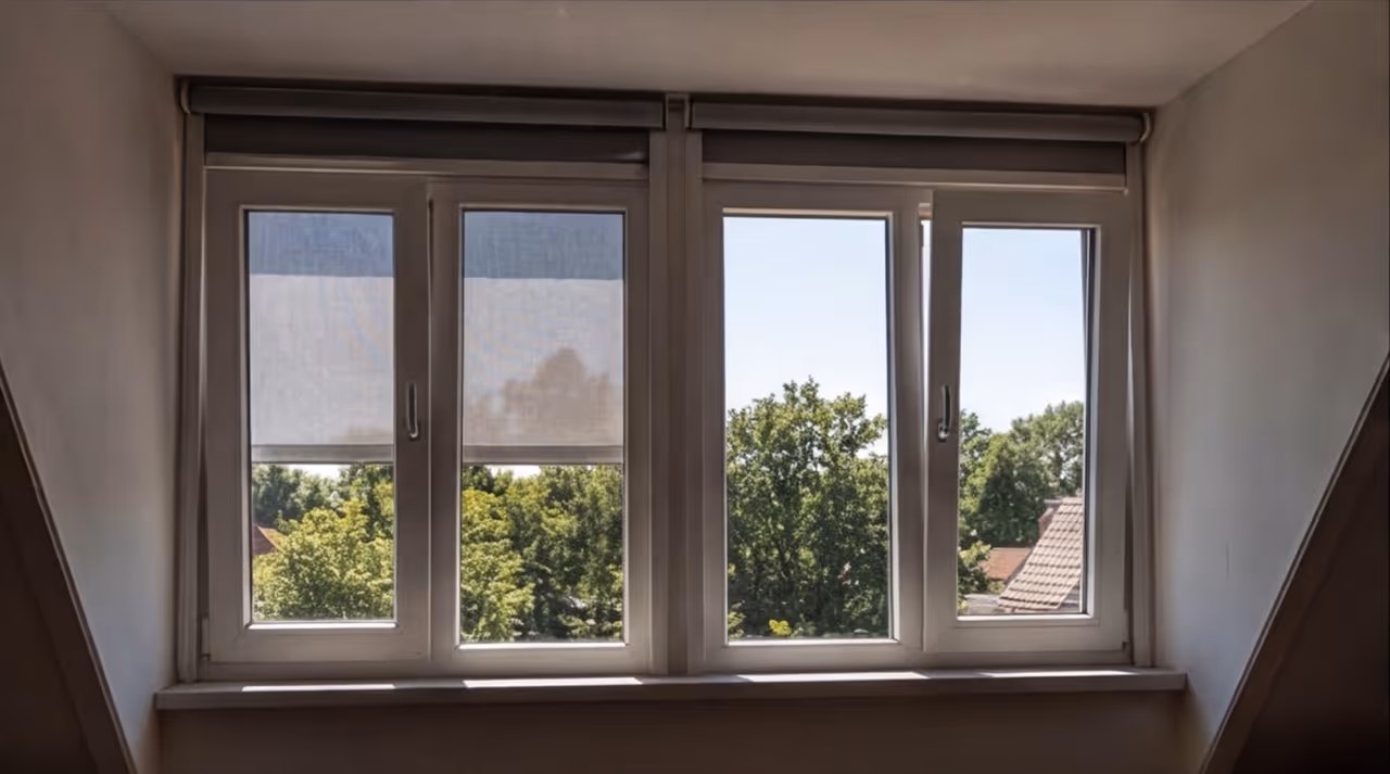 Vierdelig raam met rolluiken en uitzicht op groene bomen en daken buiten.