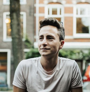 Jonge man met kort haar en wit t-shirt zit buiten met een wazige achtergrond van gebouwen.
