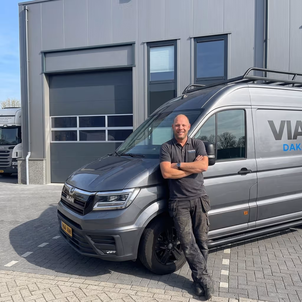 Man met gekruiste armen leunt tegen een grijze bestelwagen geparkeerd voor een bedrijfsgebouw.