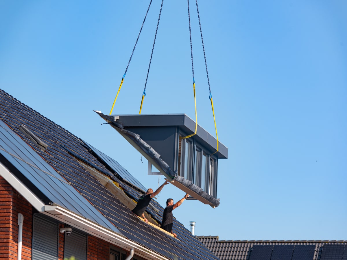 Twee mannen begeleiden een groot raam dat met kettingen aan een kraan wordt opgetild naar het dak van een huis.